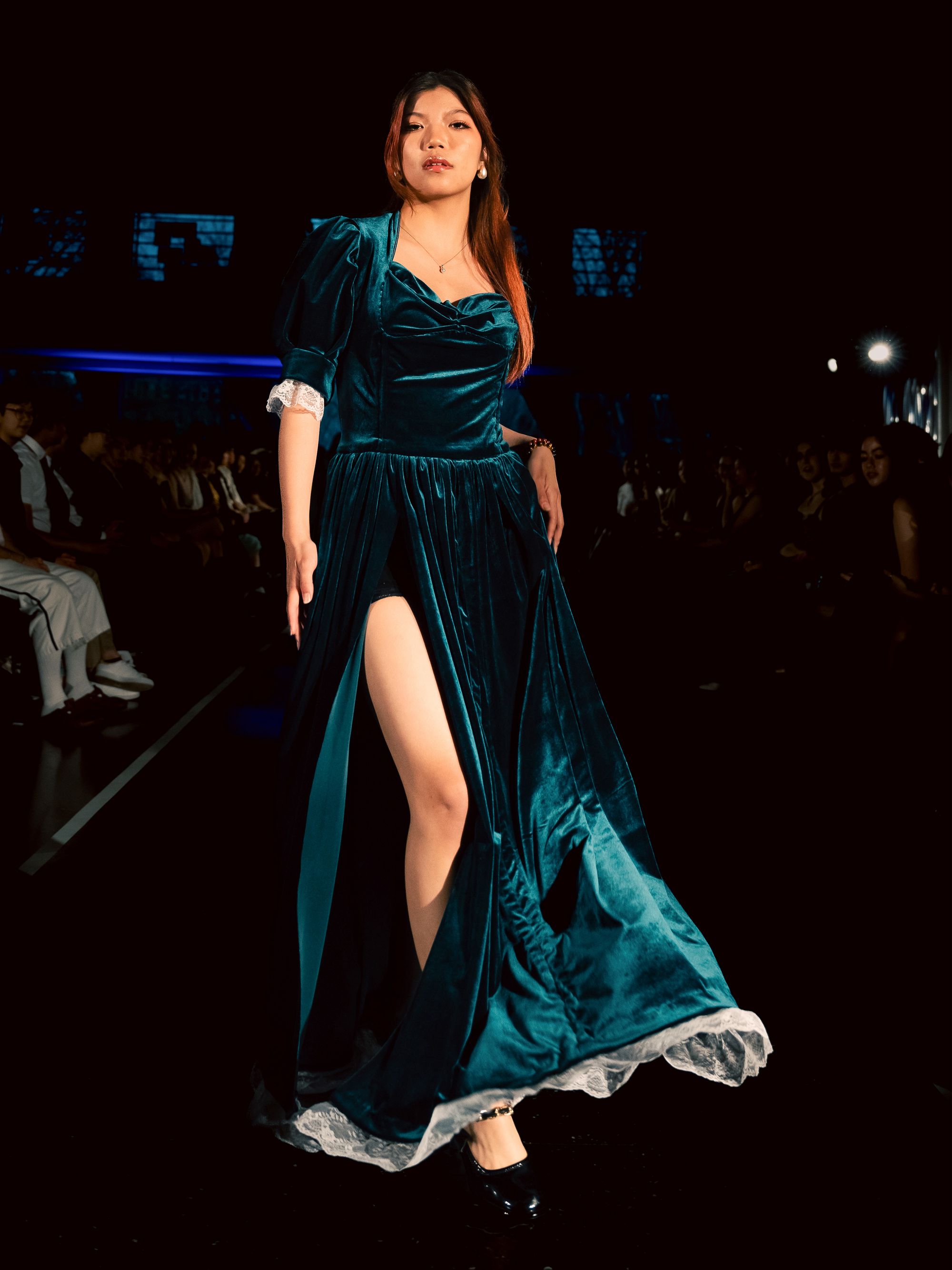Model wears a deep teal velvet gown with puffed sleeves, a high slit revealing black shorts, and a subtle lace trim at the hem. She strides down the runway under a spotlight.
